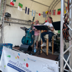 Zwei Musiker auf Bühne unter Zeltdach, einer spielt Kontrabass, der andere Gitarre und singt, dahinter internationale Flaggen