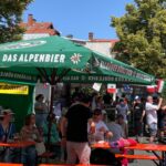 Menschen sitzen und stehen an roten Biertischgarnituren unter grünem Sonnenschirm