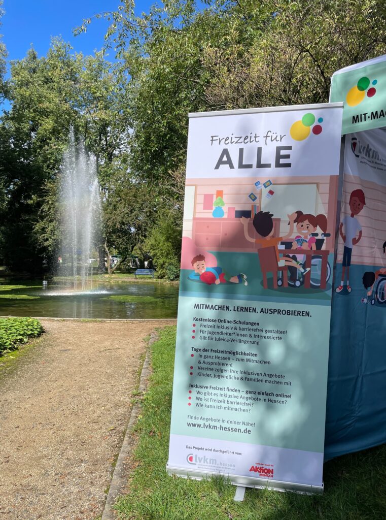 Ein Banner mit dem Text 'Freizeit für ALLE' und Illustrationen von Kindern, die an einem Tisch spielen. Im Hintergrund ein Brunnen und Bäume.