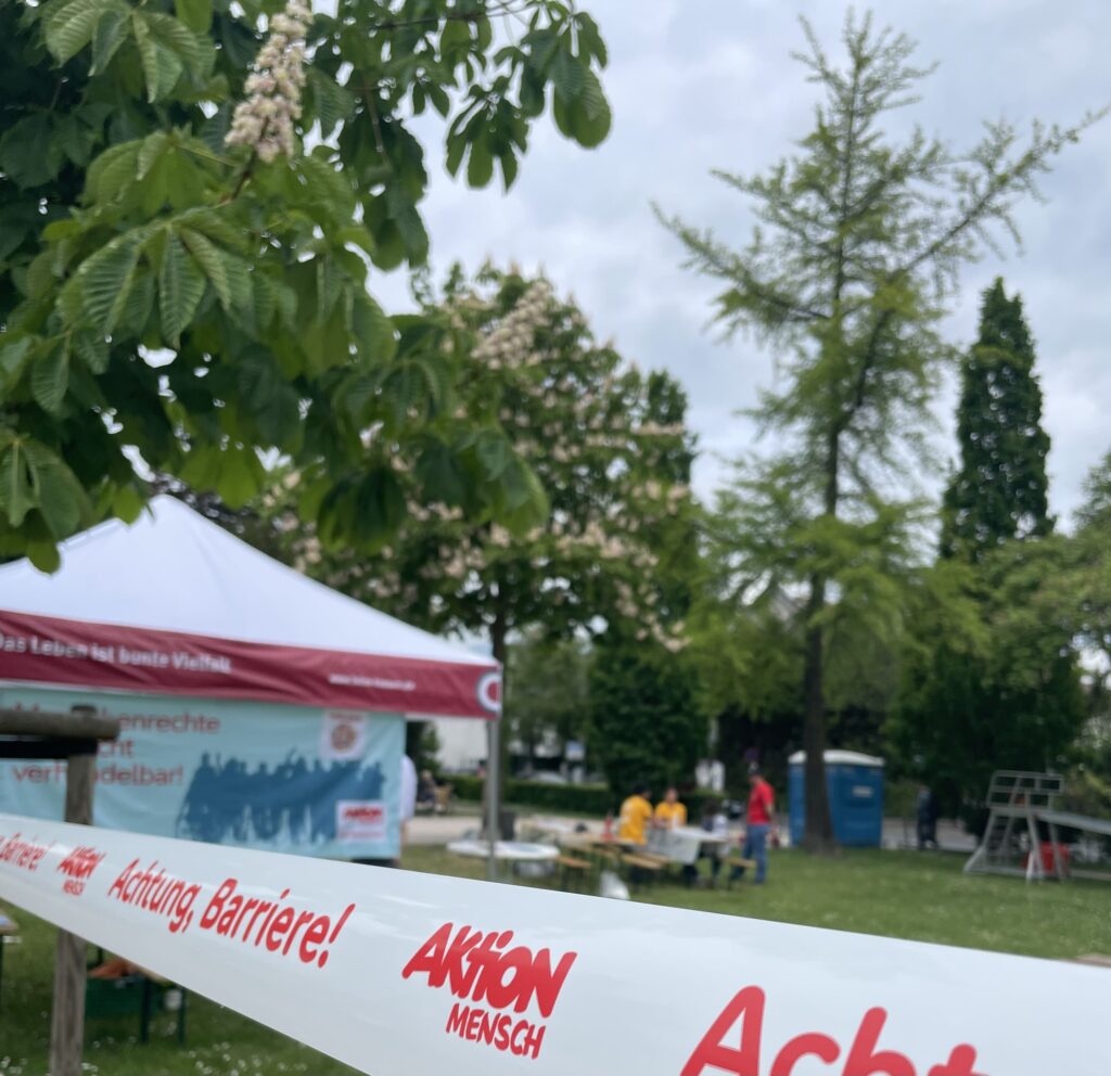 Absperrband mit rotem Aufdruck 'Achtung Barriere!' und 'Aktion Mensch' vor einem Pavillon in einem Park
