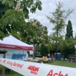 Absperrband mit rotem Aufdruck 'Achtung Barriere!' und 'Aktion Mensch' vor einem Pavillon und Bäumen in einem Park