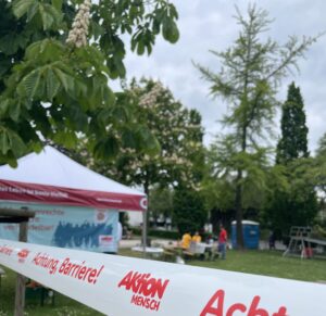 Absperrband mit rotem Aufdruck 'Achtung Barriere!' und 'Aktion Mensch' vor einem Pavillon in einem Park