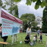 Menschen stehen und sitzen auf einer Wiese vor einem Pavillon mit Bannern, auf einem Banner steht 'Menschenrechte sind nicht verhandelbar!'