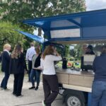 Menschen stehen an einem blauen mobilen Caféwagen unter einem blühenden Baum