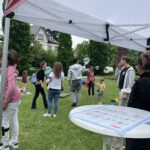 Menschen stehen auf einer Wiese unter einem weißen Pavillon mit rotem Rand und verteilen sich um einen runden Tisch mit Informationsmaterial