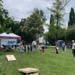 Gruppe von Menschen, darunter Kinder, auf einer Wiese mit verschiedenen Spielangeboten und einem Pavillon mit einem Banner im Hintergrund