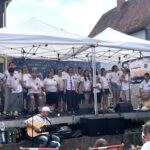 Der Chor steht auf der Bühne und singt. Im Vordergrund spielt eine Person Gitarre.