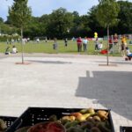 Kinder und Erwachsene bewegen sich auf einem Sportplatz, im Vordergrund steht eine Kiste mit Obst.