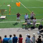 Mehrere Kinder und Erwachsene sitzen und stehen am Rand eines Sportplatzes mit aufgebautem Parcours und bunten Luftballons.