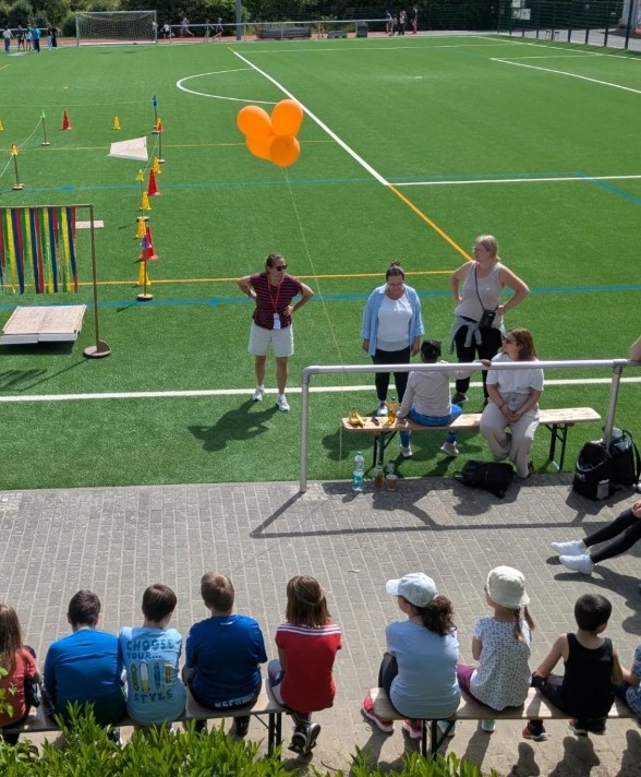 Mehrere Kinder und Erwachsene sitzen und stehen am Rand eines Sportplatzes mit aufgebautem Parcours und bunten Luftballons.