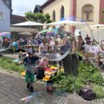 Kind mit ausgestreckten Armen spielt vor einer Gruppe sitzender Menschen mit großen Seifenblasen im Freien.