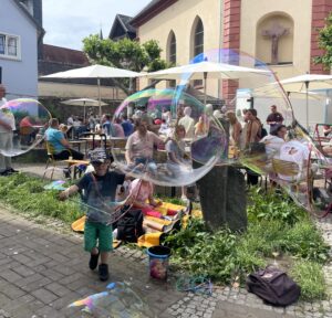 Kind mit ausgestreckten Armen spielt vor einer Gruppe sitzender Menschen mit großen Seifenblasen im Freien.