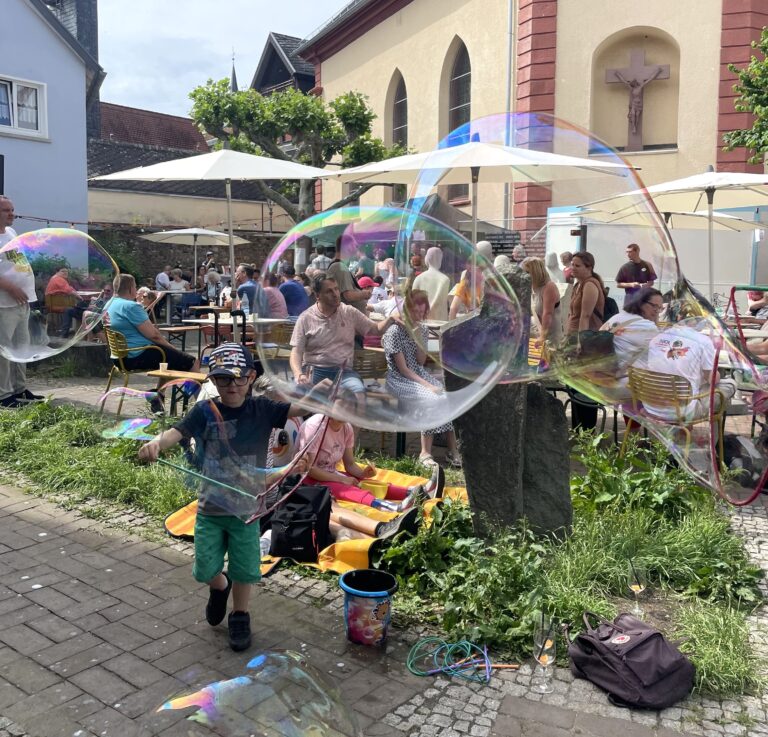 Kind mit ausgestreckten Armen spielt vor einer Gruppe sitzender Menschen mit großen Seifenblasen im Freien.