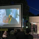 Blick auf die Leinwand, rechts daneben stehen zwei Gebärdensprachdolmetscherinnen