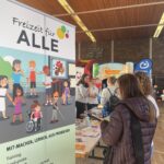 Mehrere Personen stehen an einem Infostand in einer Halle neben einem Rollup mit der Aufschrift 'Freizeit für ALLE'