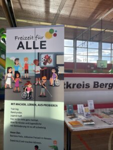 Rollup mit gezeichneten Kindern und Erwachsenen mit und ohne Behinderung, daneben Infotisch in Sporthalle