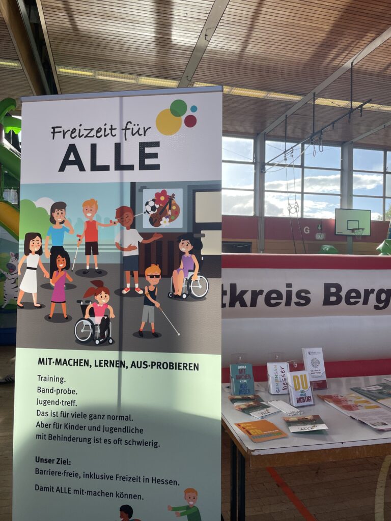 Rollup mit gezeichneten Kindern und Erwachsenen mit und ohne Behinderung, daneben Infotisch in Sporthalle