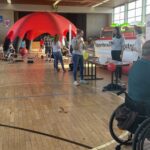 Mehrere Menschen in einer Sporthalle, darunter eine Person im Rollstuhl im Vordergrund und ein großer roter Pavillon im Hintergrund.