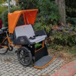 Dreirädriges Lastenfahrrad mit orangefarbenem Dach und grauer Sitzfläche auf einem gepflasterten Weg vor Sträuchern