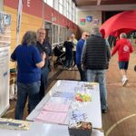 Mehrere Erwachsene unterhalten sich an einem Infostand mit Flyern, Broschüren und kleinen Werbegeschenken in einer Sporthalle.