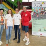 Vier Personen stehen nebeneinander in einer Halle vor einem Plakat mit der Überschrift 'Freizeit für ALLE'